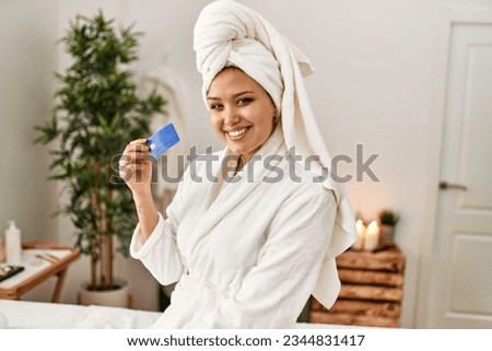 Young beautiful hispanic woman wearing bathrobe holding credit card at beauty salon