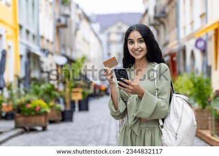 Beautiful young Indian woman using phone and credit card. Standing outside on a city street and smiling at the camera. Royalty-Free Stock Photo #2344832117