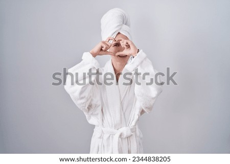 Blonde caucasian woman wearing bathrobe doing heart shape with hand and fingers smiling looking through sign 