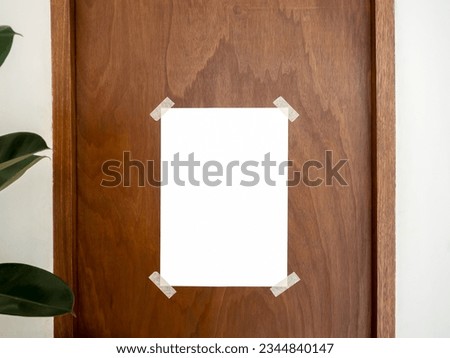 Mockup empty white vertical poster taped on vintage brown wooden door and white wall background near green leaves in cafe, minimal style. Blank poster template with tape on the corner, for advertise.