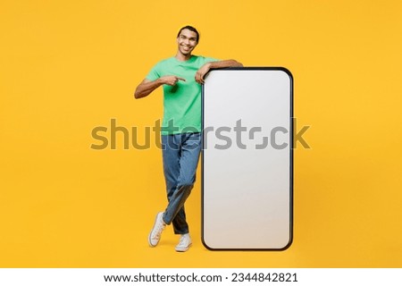 Full body young man he wearing casual clothes green t-shirt hat point index finger on big huge blank screen mobile cell phone smartphone with area isolated on plain yellow background studio portrait