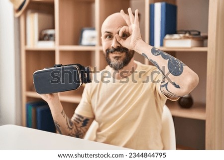 Young hispanic man wearing virtual reality glasses smiling happy doing ok sign with hand on eye looking through fingers 