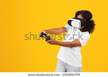 Positive teenage black pupil girl in white t-shirt, VR glasses with joystick enjoys spare time, playing online game isolated on yellow background, studio. Gadget for fun, lifestyle