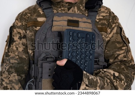 Soldier in grey bulletproof jacket with icon of ukrainian flag and keyboard in arm in black glove.