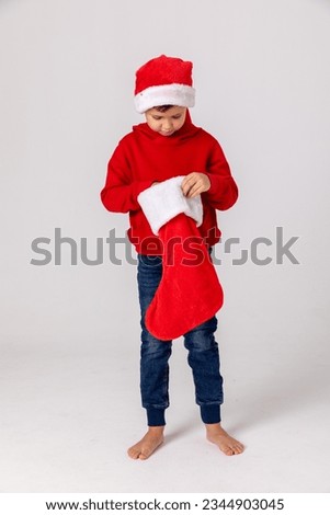 Kids opening Christmas presents. Child little boy in Santa red hat searching for candy and gifts in Christmas red sock on winter morning on white background. Winter holidays concept. Happy family time