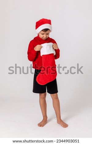 Kids opening Christmas presents. Child little boy in Santa red hat searching for candy and gifts in Christmas red sock on winter morning on white background. Winter holidays concept. Happy family time