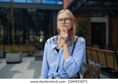 Thoughtful Caucasian woman keeps hand on chin looks pensive above poses against business build background blank copy space for your advertising content thinks about future. High quality photo