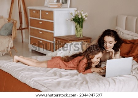 Mother and daughter use laptop on bed on vacation at home. The concept of using online services for online shopping, watching video content or cartoons on streaming services.