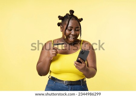 Smiling happy african american plus size model holding mobile phone and credit card paying online shopping isolated on yellow background. Beautiful female in casual clothes using smartphone