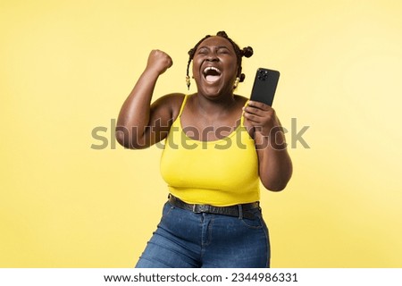 Overjoyed African American woman holding mobile phone with closed eyes making winning gesture isolated on yellow background. Beautiful plus size female with stylish hairstyle online shopping concept