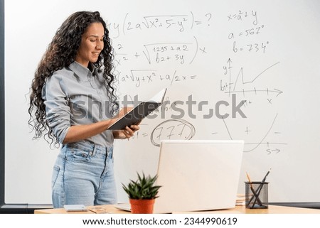 Educational Concept, E-Learning. Portrait of Middle Eastern female Math teacher standing near whiteboard with Math Equations Graphics, holding and reading notes, having online video call on laptop