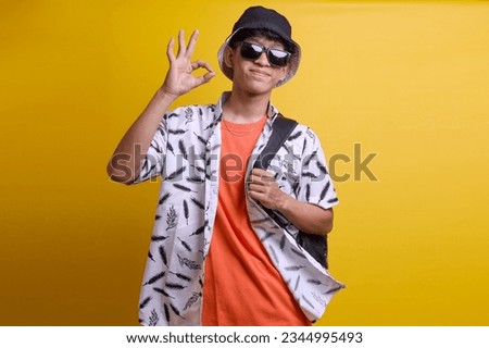 Young asian traveler man in casual clothes and sunglasses, showing ok sign and smiling over isolated yellow background. 