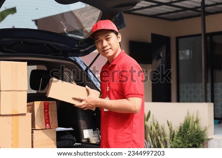 Delivery man holding cardboard box near his delivery car and looking at camera smiling.   