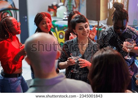 Smiling woman drinking alcohol with group of friends and enjoying disco party in nightclub. Cheerful young people holding beverage glasses, saying toast and clubbing on dancefloor