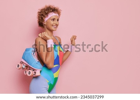 Happy curly girl dressed in colorful leotard stands isolated on pink background with pair of roller skates, sport lifestyle concept, copy space