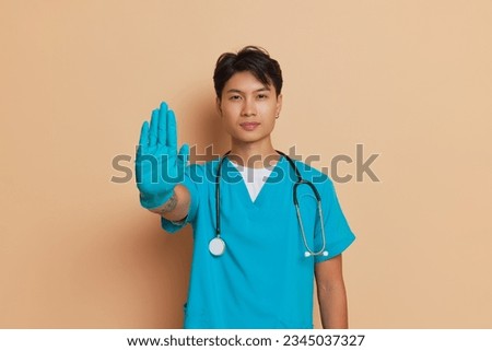 Young man in turquoise medical clothes and latex gloves shows stop gesture with his hand, professional people concept, copy space