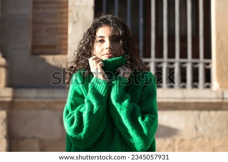 Portrait of beautiful young brunette woman with curly hair and green woollen coat covering her face with the collar of the coat for the cold. The woman is happy and looks at the camera for the photo.