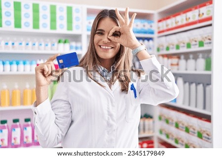 Hispanic young woman working at pharmacy drugstore holding credit card smiling happy doing ok sign with hand on eye looking through fingers 