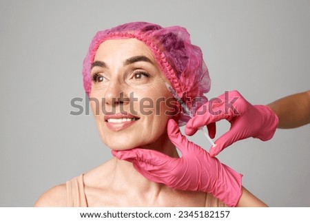 Middle-aged woman in pink caps for cosmetic treatments doing beauty injection. Hands in pink gloves hold a syringe. Face lift. Beauty, cosmetology, anti-aging, skin care, injections concept. Royalty-Free Stock Photo #2345182157