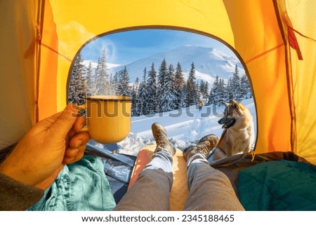 Hot drink in the hand and wonderful view of snowy forest through the open entrance to the tent. The beauty of a romantic hike and camping accompanied by a dog.