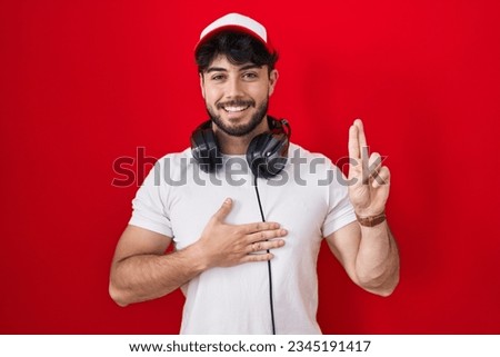 Hispanic man with beard wearing gamer hat and headphones smiling swearing with hand on chest and fingers up, making a loyalty promise oath 