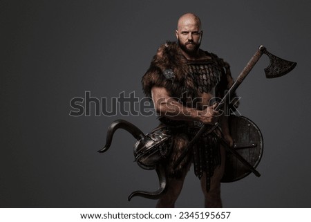 A rugged, bald, bearded Viking dressed in fur and light armor, with a helmet attached to his belt, holding an axe and shield against a gray background