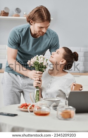 Vertical portrait of smiling young man giving flowers to wife as surprise in morning