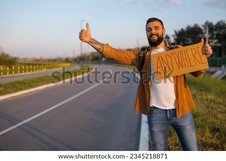 Man is hitchhiking on roadside trying to stop car. He is holding cardboard with inscription.