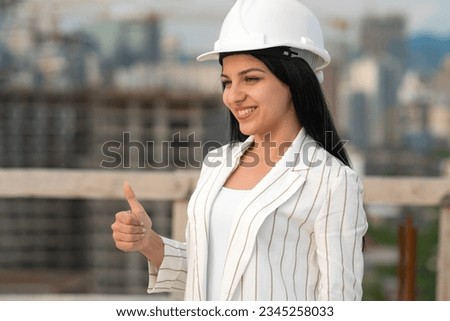 Young beautiful smiling woman in a white helmet shows a thumbs up sign, OK on a construction site. Concept of the profession of architect, engineer, manager, surveyor