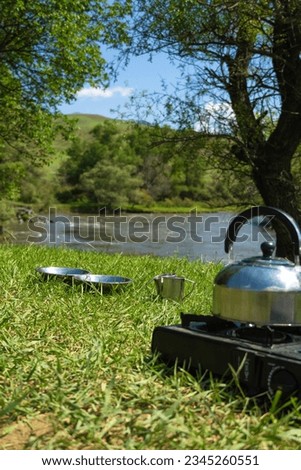 Close-up of aluminum plates, mugs, a camping gas burner with a kettle stand on the green grass against the background of a mountain river and trees on a salty day. Cooking while traveling by car.