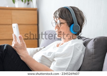 Senior woman listening to music on mobile phone wearing wireless headphones sitting on couch relaxing at home. Elder mature woman listens audiobook online or using musical application indoor