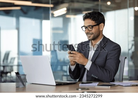 Successful experienced businessman using phone while sitting at workplace, hispanic smiling happy with achievement results holding phone, reading online using app on smartphone. Royalty-Free Stock Photo #2345272299