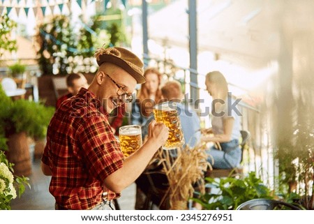 Close-up of young man in checkered shirt and fedora hat drinking beer. Blurred people in background. Sunny day. Concept of oktoberfest, traditional taste, friendship, leisure time, enjoyment