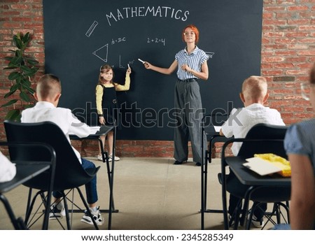 Young woman, teacher standing by blackboard with little girl, child and teaching math at school. Attentive kids listening. Concept of school, education, childhood, knowledge, lifestyle