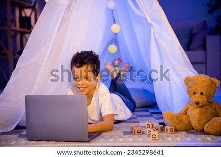 Photo of joyful adorable boy have fun evening weekend watch video on netbook indoors in night playroom