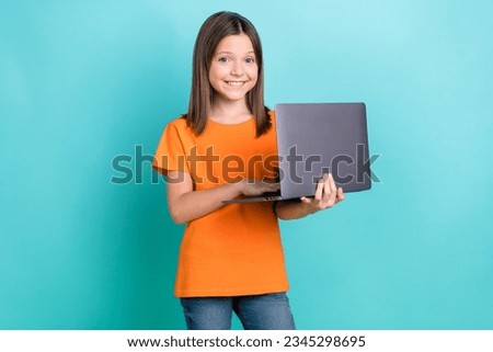 Photo of smart clever positive little girl wear orange stylish t-shirt holding laptop typing email isolated on teal color background