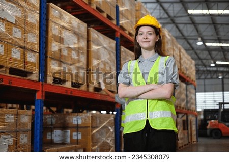 Portrait of smiling female factory worker with her arms crossed confidently in the process of working in warehouse.Industrial engineer wearing hard hat and safety vest stand beside inventory shelves.