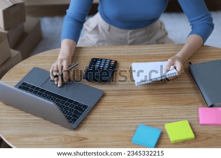 Asian woman starting a small business at home working with parcel boxes and calculating revenue and profit from sales.