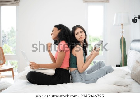 Two young women sitting on bed in bedroom and using laptop and tablet for play the game.