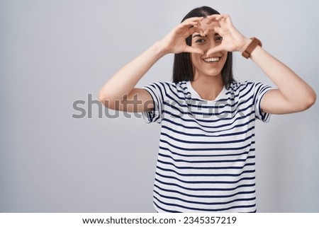 Young brunette woman wearing striped t shirt doing heart shape with hand and fingers smiling looking through sign 