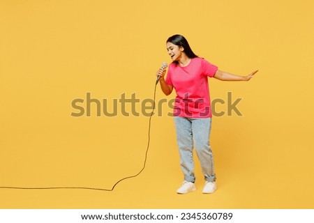Full body singer young happy Indian woman wearing pink t-shirt casual clothes sing song in microphone at karaoke club stage isolated on plain yellow wall background studio portrait. Lifestyle concept Royalty-Free Stock Photo #2345360789