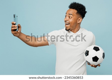 Young man fan wear basic t-shirt do selfie shot on mobile cell phone cheer up support football sport team hold in hand soccer ball watch tv live stream isolated on plain pastel blue color background