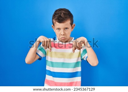 Young caucasian kid standing over blue background pointing down looking sad and upset, indicating direction with fingers, unhappy and depressed. 