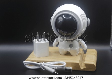 White astronaut doll with a power plug on a black background.  A white tuk tuk stands.