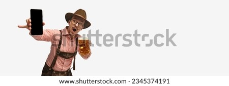 Wow, surprised. Young mustache man, waiter in traditional Bavarian ot German outfit show phone with blank screen. Celebration, oktoberfest, festival, trades, graphs concept. Copy space for ad. Banner