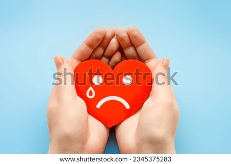 female hands hold a paper heart with a crying smiley face isolated over blue background. broken heart love rejection. hidden narcissist psychology concept