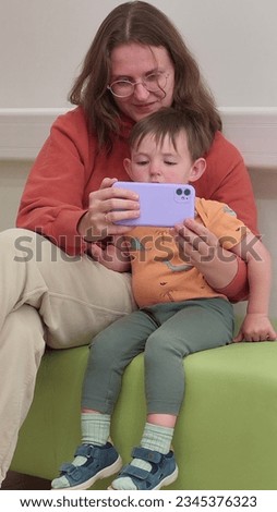 The mother and her baby are in the hospital waiting room, reading a book on the mobile phone together while they wait for their doctor's visit. Kid boy aged two years (two-year-old)