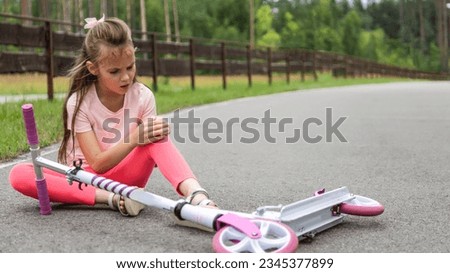 Little girl sitting on the ground after she fell while riding her scooter at summer park Royalty-Free Stock Photo #2345377899