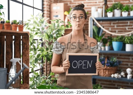 Hispanic woman working at florist holding open sign making fish face with mouth and squinting eyes, crazy and comical. 