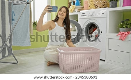 Young beautiful hispanic woman washing clothes make selfie by smartphone at laundry room